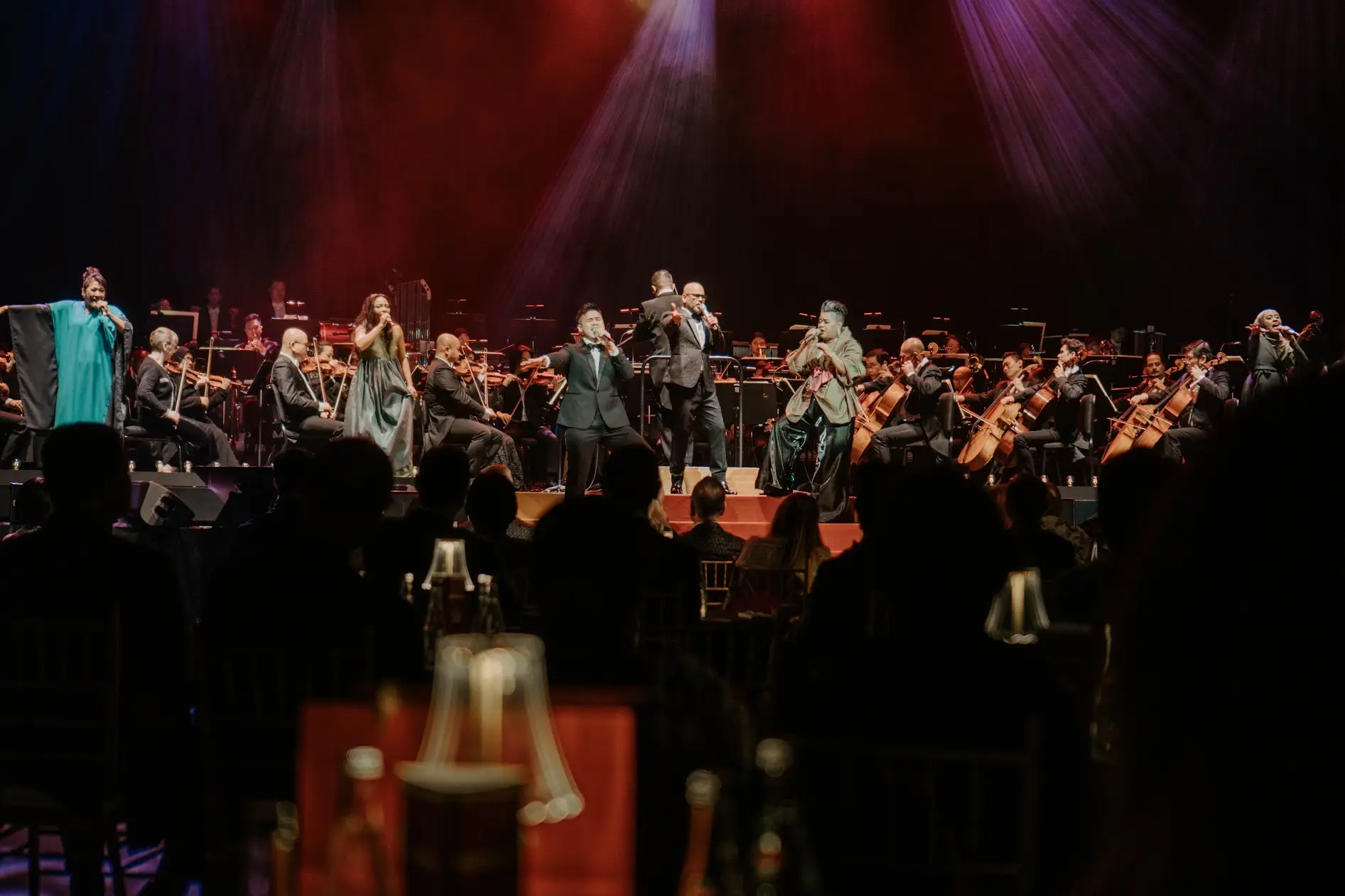 The orchestra and singers close to, lit warm red and violet, seen past the heads of the audience.