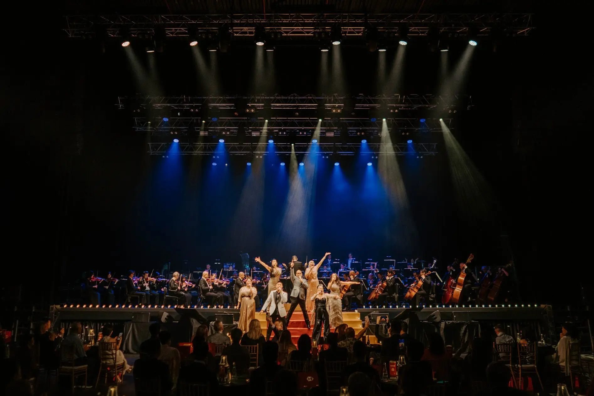 Performers with their arms raised at the curtain call, white and blue beams crossing overhead.