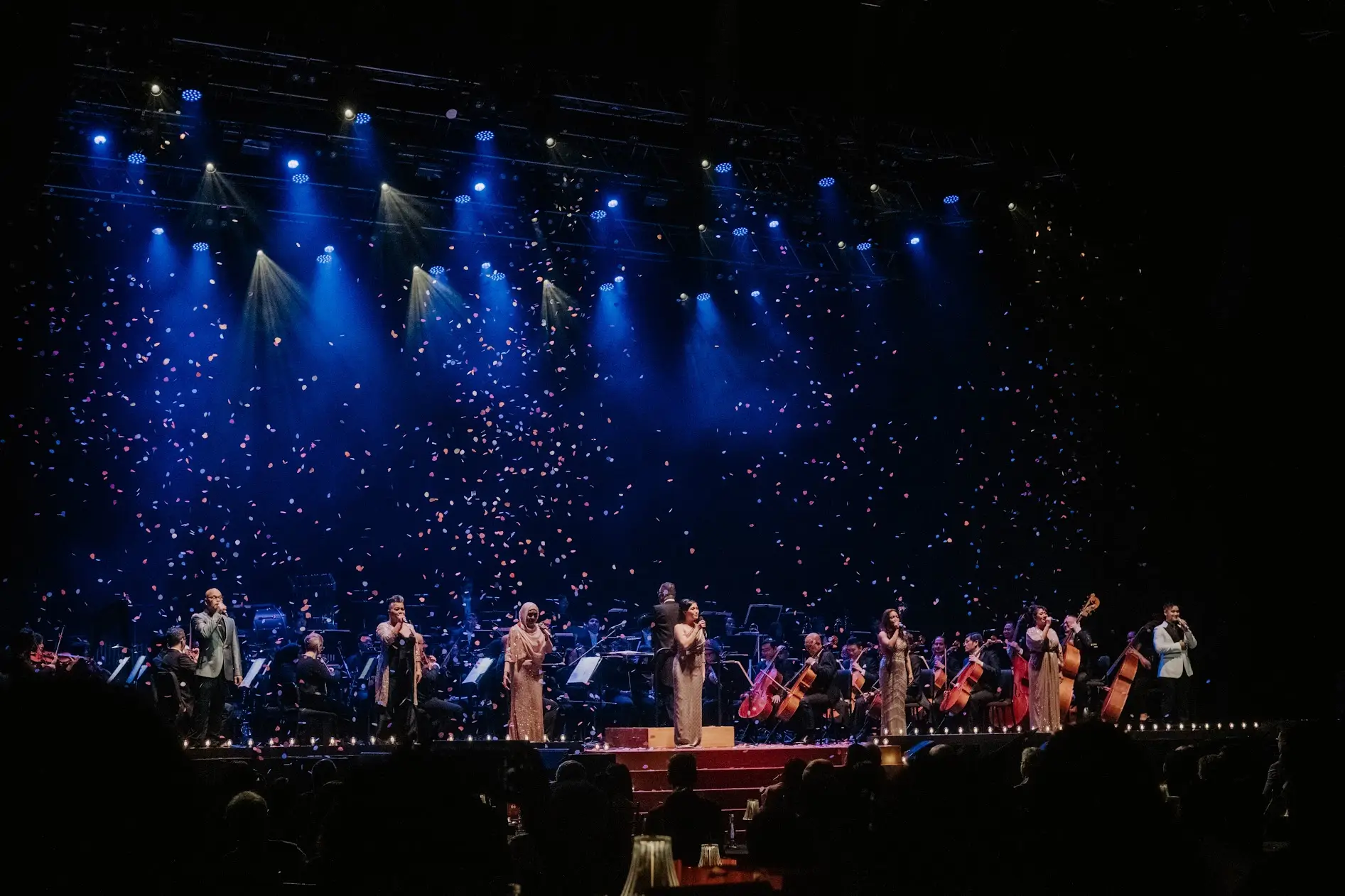 Singers downstage with the orchestra behind them, blue beams raking across the hall.