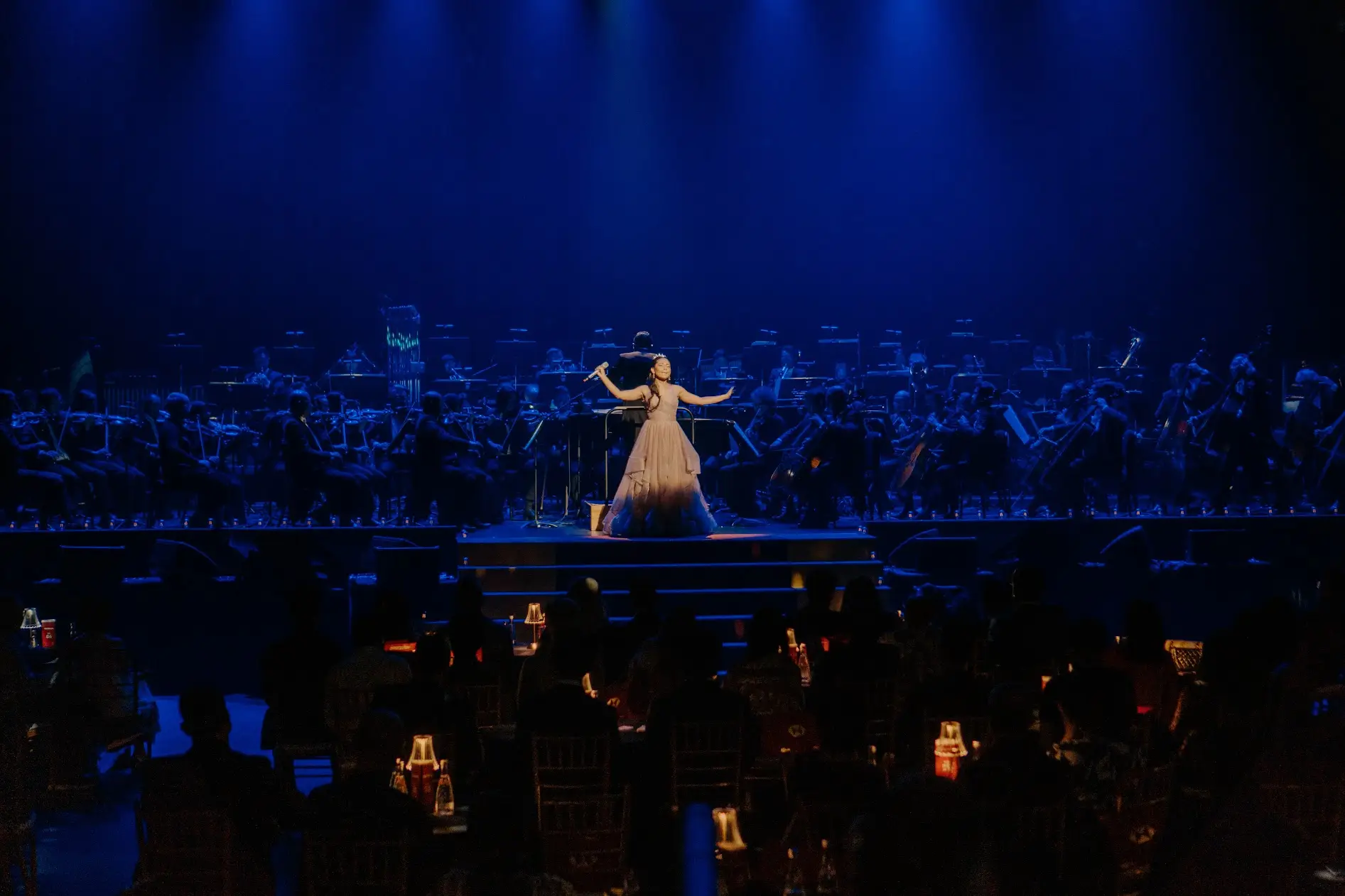 A soloist alone downstage in a pale gown, the orchestra ranked behind in blue light.